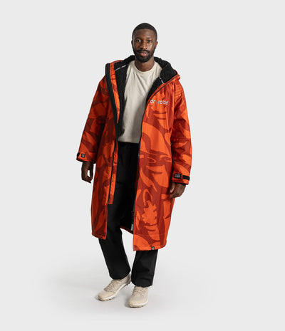 *MALE* A man modeling a long orange camouflage hooded Dryrobe with black trim, worn over a white t-shirt and black pants, on a light gray studio background.