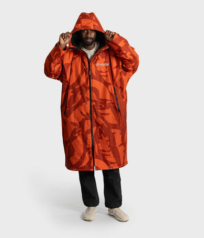 *MALE* A man wearing a long hooded Dryrobe with an orange and red abstract camouflage pattern, black pants, and beige sneakers, standing against a light gray studio background. The brand name 'dryear' is visible on the chest.