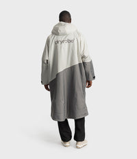 *MALE* A person shown from the back wearing a long hooded Dryrobe in off-white and grey colour blocking, paired with black pants and white sneakers, against a light grey studio background.