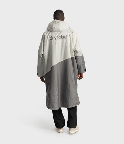 *MALE* A person shown from the back wearing a long hooded Dryrobe in off-white and grey colour blocking, paired with black pants and white sneakers, against a light grey studio background.