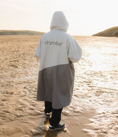 *MALE* A person standing on a sandy beach at sunset wearing a colour-blocked hooded Dryrobe with 'dryrobe' printed on the back. The Dryrobe is white on the upper half and grey on the lower half.