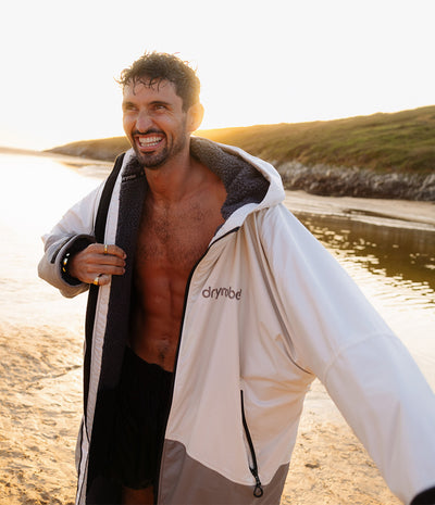 *MALE* A smiling man on a beach at sunset wearing an open Dryrobe hooded jacket, showing the dark fleece-lined interior and the light outer shell. The background shows sand, water, and grassy dunes.