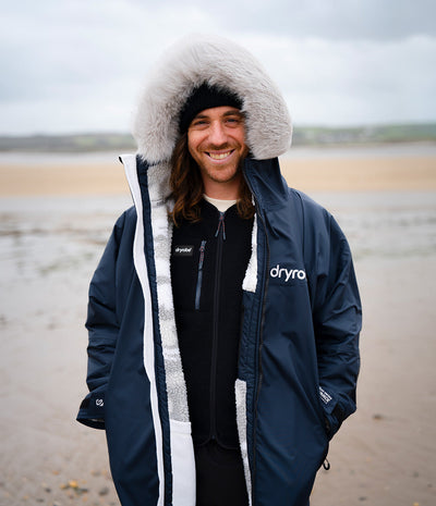 *MALE* A smiling man wearing a navy blue Dryrobe with a white faux-fur trimmed hood, standing on a sandy beach under an overcast sky.