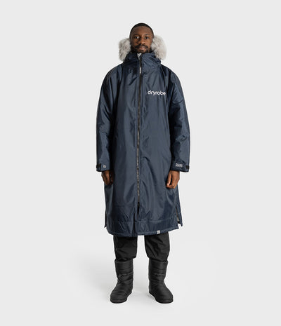 *MALE* A man wearing a long navy blue Dryrobe with a light gray faux-fur trimmed hood, black pants, and black boots, standing against a light gray studio background.