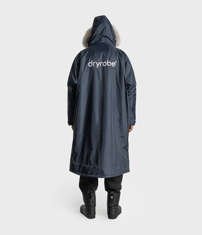 *MALE* A person shown from the back wearing a long navy hooded Dryrobe with a light fur-trimmed hood and a white dryrobe logo on the back, paired with black pants and black boots against a light gray studio background.