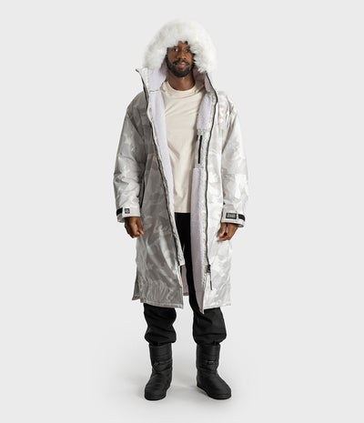 *MALE* A man wearing a long, white hooded Dryrobe with a white faux-fur trim, layered over a cream t-shirt, black pants, and black boots, shot against a light gray studio background.