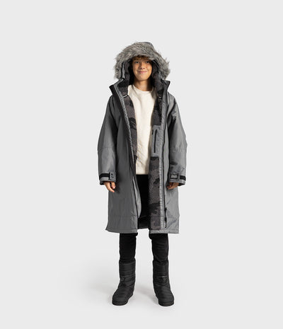 *MALE* A child wearing a long gray hooded Dryrobe with a faux-fur trimmed hood, layered over a cream sweater, black pants, and black boots, standing against a light gray studio background.