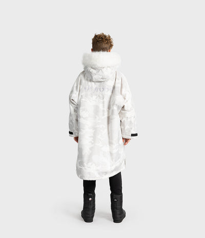 *MALE* A child viewed from the back wearing a long white hooded Dryrobe with a subtle tonal pattern, a large white faux-fur trimmed hood, black pants, and black boots against a light gray studio background.