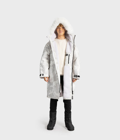 *MALE* A child wearing a long white and gray camouflage hooded Dryrobe with a fluffy white faux-fur trim, layered over a cream sweatshirt, black pants, and black boots, photographed against a light gray studio background.