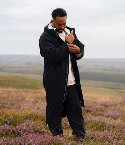 *MALE* A man wearing a long black hooded raincoat over a white shirt and black trousers, standing in a field of purple heather with rolling hills in the background under an overcast sky.