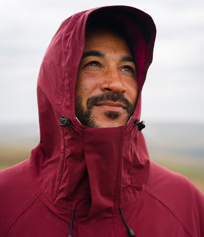 *MALE* A close-up shot of a man wearing a burgundy hooded rain jacket, with the hood up and his face partially covered, set against a soft-focus outdoor background.