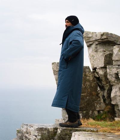 *MALE* A man wearing a long navy blue hooded coat, black beanie, and hiking boots stands on a rocky cliffside overlooking a misty body of water under an overcast sky.