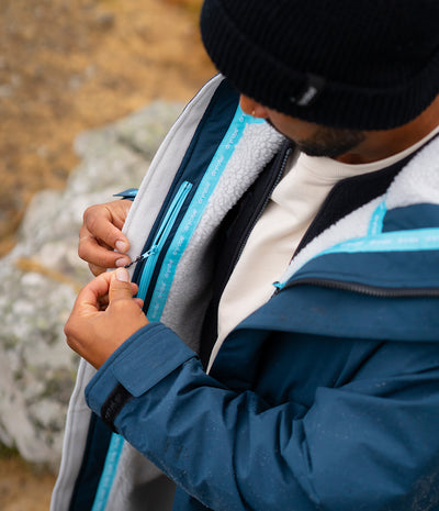 *MALE* A close-up lifestyle shot of a man wearing a blue and gray fleece-lined jacket, adjusting the zipper on the chest. The jacket features a bright blue zipper track with white text that reads 'TERRAFORM'.