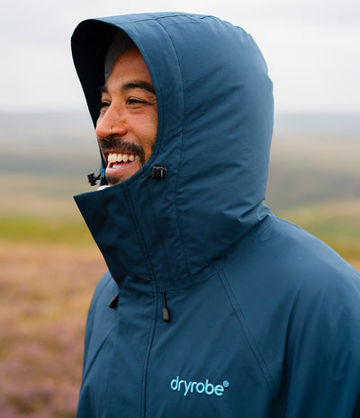 *MALE* A man wearing a navy blue Dryrobe hooded rain jacket, smiling outdoors against a blurred natural landscape background.