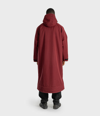 *MALE* A person shown from the back wearing a long hooded coat in a deep burgundy or maroon color, paired with black pants and dark shoes, against a plain light gray studio background.