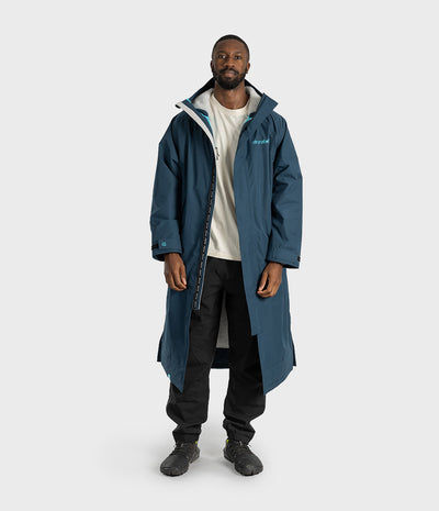 *MALE* A man wearing a long, dark teal-blue hooded raincoat with a small light blue 'Dryrobe' logo on the left chest, paired with black pants and dark boots, standing against a light grey studio background.