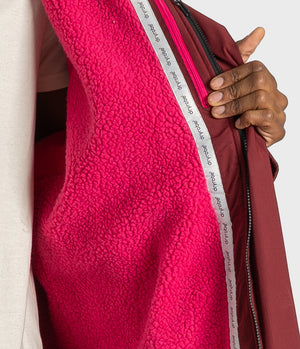 Close-up of a person holding open a burgundy zip jacket to show a bright pink sherpa fleece lining. A white zipper tape with the brand name 'arbitro' is visible.