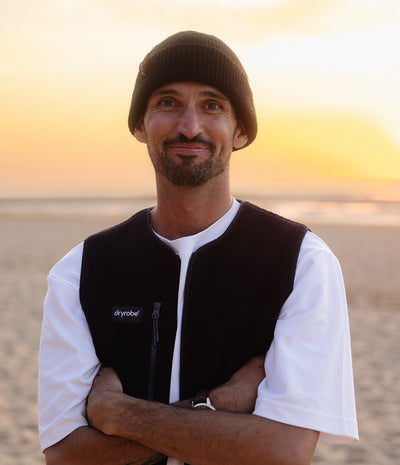 A man wearing a black knit beanie, a black Dryobot fleece vest over a white t-shirt, standing on a sandy beach at sunset.