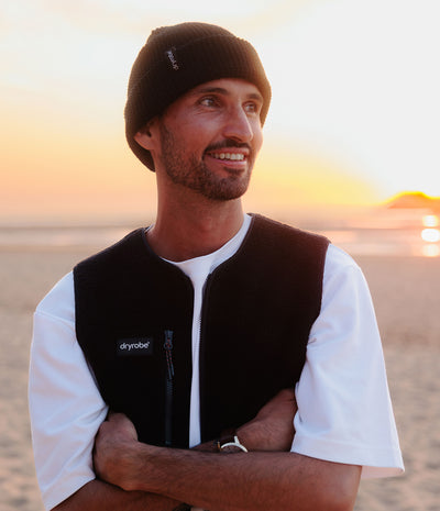 A man wearing a black knit beanie and a black Drymobile fleece vest over a white t-shirt, standing on a beach at sunset.