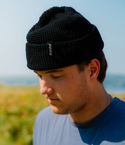 A man wearing a black knit beanie with a small white 'dryrope' logo tag, photographed outdoors against a blurred blue sky and grassy background.