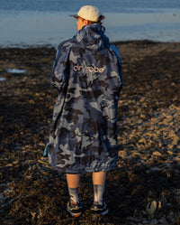 **MALE** Man stood with back to the camera on a beach, wearing dryrobe Advance Long Sleeve changing poncho in Blue Camo Blue