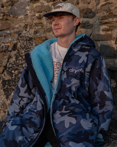 **MALE** Man sat against a wall wearing dryrobe Advance Long Sleeve changing robe in Blue Camo Blue