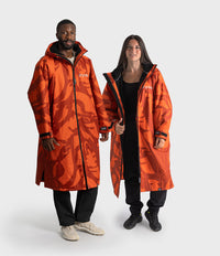 A man and a woman modeling matching bright orange waterproof Dryrobe with a darker orange abstract camouflage pattern, black zippers, and white Arc'teryx logos on the chest, set against a plain light gray studio background.