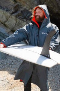Man in a gray hooded Dryrobe holding a surfboard on a rocky beach.