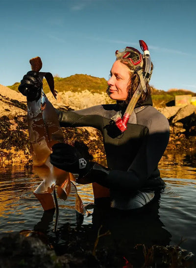 Person in scuba gear holding a small figurine by a body of water with a scenic background.