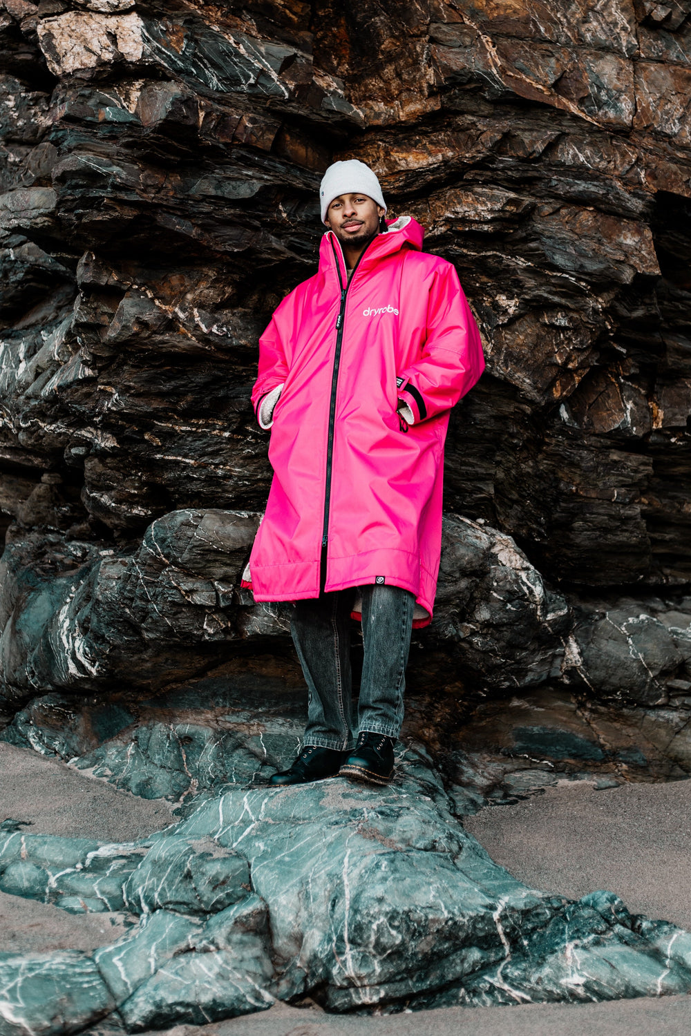 A person wearing a bright pink hooded dryrobe, a white beanie, and dark pants stands on a rocky outcrop in front of a large dark rock face.