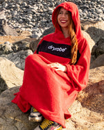 Woman sat on rocks on the beach wearing Organic Towel dryrobe Remix in Red Black with hood up 
