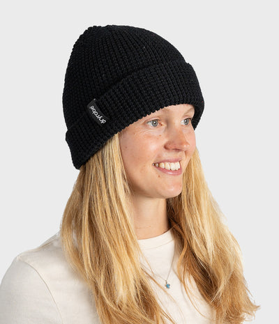 A smiling blonde woman wearing a black ribbed knit beanie with a small white 'Dryrobe' brand tag on the side, photographed against a light gray studio background.