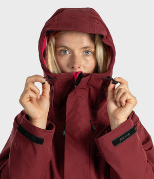 A close-up of a woman wearing a hooded technical jacket in a deep burgundy color, holding the hood up with both hands against a light gray studio background.