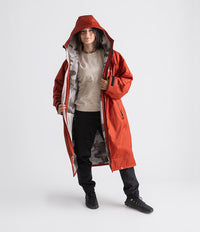 A woman modeling a long hooded red waterproof jacket with a gray and white patterned lining, worn over a beige sweatshirt, black pants, and black boots, against a light gray studio background.