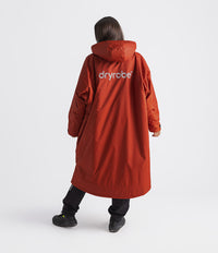 A person shown from the back wearing a long hooded orange-red dryrobe jacket with a white 'dryrobe' logo on the back, paired with black pants and black shoes, against a light gray studio background.