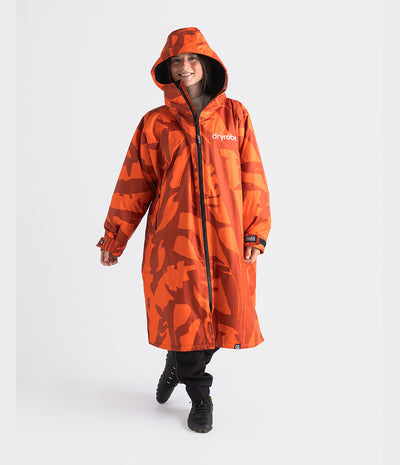 A woman wearing a bright orange hooded Dryrobe with a dark abstract camouflage pattern and the brand name 'dryvoda' on the chest, standing against a light gray studio background.