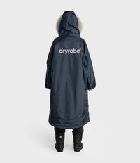 A person shown from the back wearing a long navy hooded Dryrobe with a light fur-trimmed hood and a white 'dryrobe' logo on the back, paired with black pants and boots, against a light gray studio background.