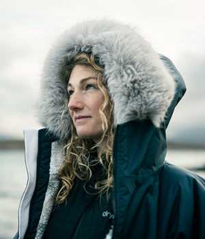 A woman wearing a dark navy winter Dryrobe with a large gray faux-fur trimmed hood, photographed outdoors against an overcast sky and distant water.