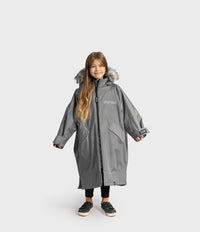 A young girl wearing a long gray Dryrobe with faux fur trim, standing against a light gray studio background.
