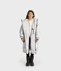 A child model wearing a long, light gray camouflage-patterned hooded Dryrobe with a white sherpa lining, styled over a cream sweater, black pants, and black boots, set against a plain light gray studio background.