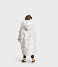 A child viewed from behind wearing a long white camouflage Dryrobe with a fluffy white faux-fur hood trim, black leggings, and black slip-on shoes, against a light gray studio background.