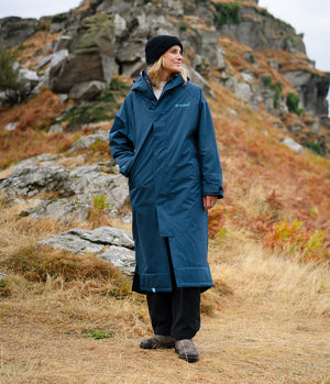 A woman wearing a long navy blue waterproof coat, black beanie, and black trousers stands outdoors in a rocky, grassy mountain setting. The brand name 'dryrobe' is visible on the left chest of the coat.