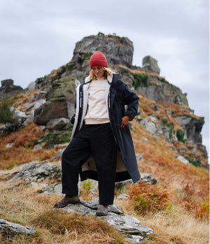A person standing outdoors on rocky grassy terrain in front of a large rock formation under an overcast sky, wearing a long dark coat over a cream top and dark trousers, with a red beanie.