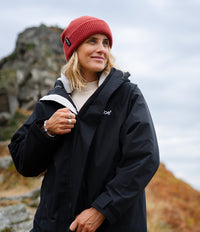 A woman wearing a black JBE waterproof jacket and a red knit beanie stands outdoors against a rocky, overcast sky background.