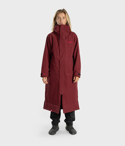 A woman modeling a long, dark red hooded technical jacket with black pants and boots, standing against a light gray studio background.