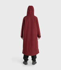 A person shown from the back wearing a long hooded coat in a deep burgundy or maroon color, paired with black pants and dark shoes, against a plain light gray studio background.
