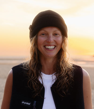 A smiling woman outdoors at sunset wearing a black knit beanie, a black sleeveless zip-front vest with a visible Dryrobe logo, and a white shirt.