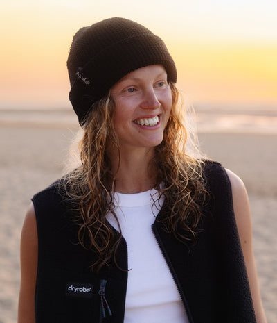 A woman smiling outdoors at sunset, wearing a black knit beanie and a black sleeveless vest with a white shirt underneath. The vest has a small white 'dryrobe' logo on the chest.
