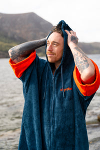 Man wearing a blue and orange hooded towel robe by a body of water with mountains in the background.