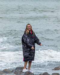 Woman stood on rocks next to the sea, wearing dryrobe Advance Long Sleeve REMIX Range change poncho in Black Camo Pink Black